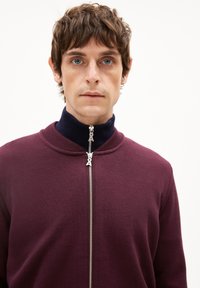 Sweatshirt zippé bordeaux avec un col en cote bleu marine, doté d'une fermeture éclair en argent et d'un tissu en maille texturée. Gros plan de la vue de face.
