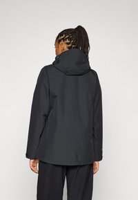 En person med fletninger har en sort hættetrøje på og ses fra bagfra, med en neutral baggrund der fremhæver tøjets design.