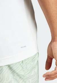 Camiseta atlética blanca con tejido texturizado, que presenta la marca HEAT.RDY en la parte inferior, combinada con pantalones cortos verdes claros que exhiben un patrón ondulado.