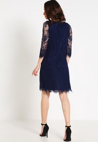 Marinblå spetsklänning med trekvartsärmar i genomskinligt material, scalloped fåll och intrikata blommönster. Svarta klackar fulländar looken.