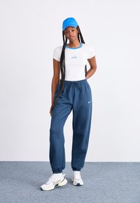 Camiseta blanca con ribetes azules y logo de "nike", combinada con pantalones jogger azul oscuro y zapatillas blancas; el modelo lleva una gorra azul a juego.