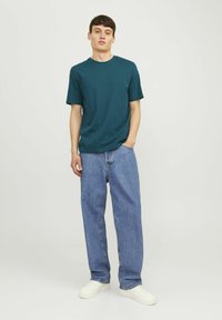 Teal katoenen T-shirt met korte mouwen, ontspannen pasvorm, gecombineerd met lichtblauwe losse jeans en witte sneakers op een neutrale achtergrond.