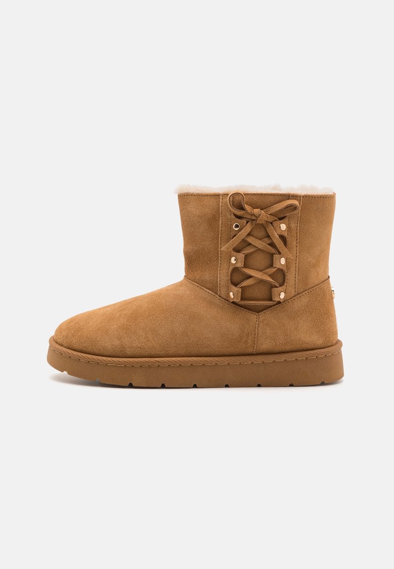 Botte d'hiver en daim beige mi-mollet avec doublure intérieure en fourrure, semelle plate et détail décoratif de laçage latéral sur fond clair uni.
