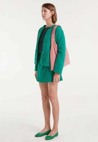 Mujer de pie, vistiendo una chaqueta verde y una falda, llevando un bolso de hombro arrugado rosa y bailarinas verdes.