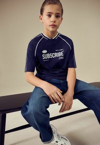 Tumesinine t-särk valgete aktsentidega ja tekstiga "SUB5CRIBE", siniste teksade ja valgete tossudega, istumas mustal pingil.