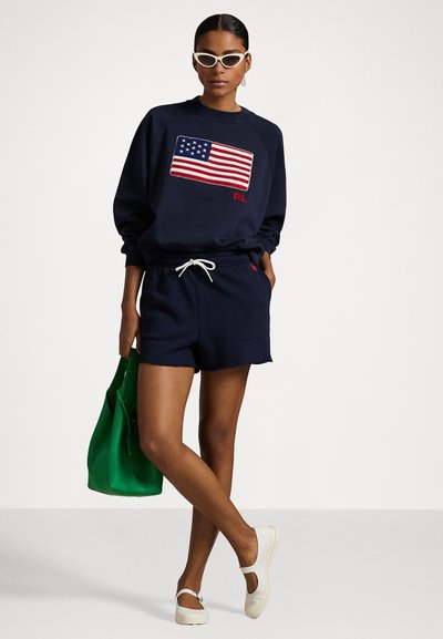 Marine sweatshirt met Amerikaanse vlag graphic, gecombineerd met marine shorts. Model houdt een groene tas vast en draagt witte schoenen. Soepele stof, comfortabele pasvorm.