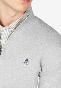 Sudadera gris con cremallera y cuello acanalado, que presenta un logotipo bordado azul en el lado izquierdo del pecho. Tejido texturizado y mangas ajustadas a la vista.