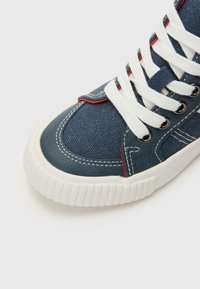 Blå lærret sneakers med hvite snøringer, røde sømmer og en teksturert gummisåle. Rund tå og kontrasterende sømdetaljer.