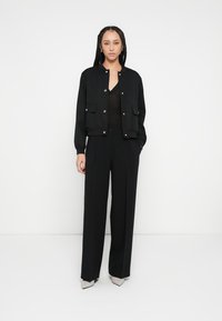 Melna bumbvedēja stila jaka ar pogu aizdari un priekšējām kabatām, valkāta virs melnas augšdaļas un saskaņota ar augstiem jostasvietas melnajiem biksēm un gaišu krāsu papēžiem.