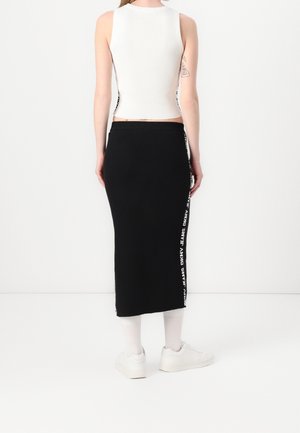 Jupe midi noire slim-fit avec texte de logo blanc sur le côté, associée à un top blanc sans manches en maille côtelée et des baskets blanches, vue de dos.
