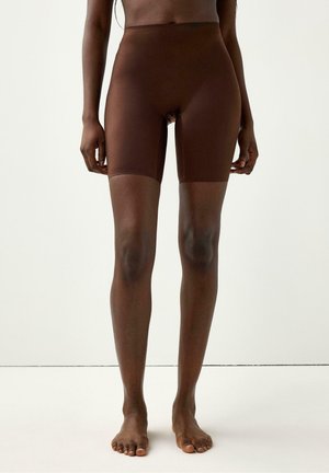 Kvinde iført mørkebrune sømløse cykelshorts med høj talje, stående barfodet mod en ensfarvet hvid baggrund.