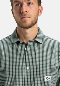 Mann mit kurzem Bart, der ein grün-schwarz kariertes Hemd mit Knopf und einer Brusttasche sowie einem DC Shoes-Logo-Patch trägt.