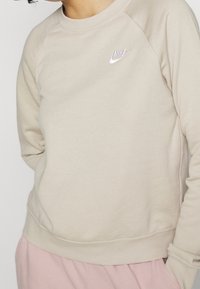 Beżowa bluza z okrągłym dekoltem i rękawami raglanowymi, z białym wyszywanym logo Nike na lewej piersi. Miła w dotyku.