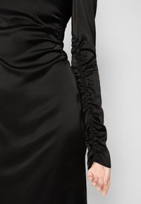 Gros plan sur une robe en satin noir avec un détail froncé le long du côté et de la manche, montrant une main partiellement couverte par le tissu.