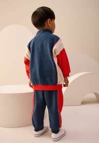 Børnesportsdragt i blå, rød og creme; har høj krave, elastiske cuffs og er lavet af blødt, struktureret stof. Komplimenteret af hvide sneakers.