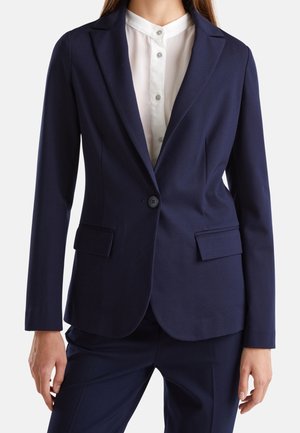 Femme portant un blazer bleu marine cintré avec des poches à rabat sur une chemise blanche à boutons, assorti à un pantalon bleu marine.