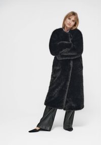 Lång svart kappa i konstpäls med heltäckande ärmar, öppen frontdesign och mjuk textur, matchad med svarta skinnbyxor och spetsiga skor.