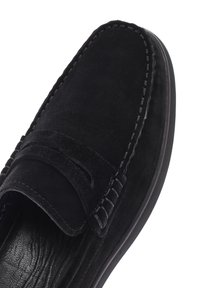 Mocasines de ante negro con un acento cosido a lo largo del empeine, suela plana y forro interior texturizado. Superficie lisa sin herrajes visibles.