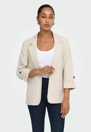 Vrouw draagt een beige blazer over een wit topje en donkere spijkerbroek, staand tegen een effen lichte achtergrond, met haar naar achteren gebonden en oorbellen in hoepelvorm.