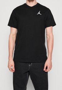 T-shirt di cotone nero con colletto rotondo e maniche corte. Presenta un logo Jumpman bianco sulla parte sinistra del petto. Indossato con jeans scuri.