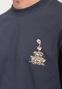 Hromada sklenic na šampaňské, do kterých se nalévá šampaňské z vrchu, potisknutá světlou barvou na tmavě modrém tričku.