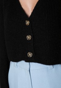 Černý pletený cardigan s hlubokým výstřihem do V, s čtyřmi kulatými průhlednými knoflíky. V kombinaci se světle modrými strukturovanými kalhotami.