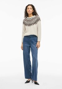 Pleteni pulover u svijetloj bež boji s uzorkom na yoke-u u tamno smeđoj i zelenoj boji. Uparen sa širokim plavim trapericama i crnim cipelama sa špicastim vrhom.