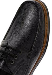 Chaussure en cuir noir avec une surface texturée, profil bas et bout rond. Comprend des lacets noirs et des surpiqûres marron contrastantes le long de la semelle.