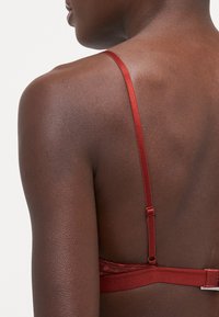 Rød blonde-bh med justerbare sateng stropper, med en teksturert bakre bredde og blomsterdetaljer langs kantene.