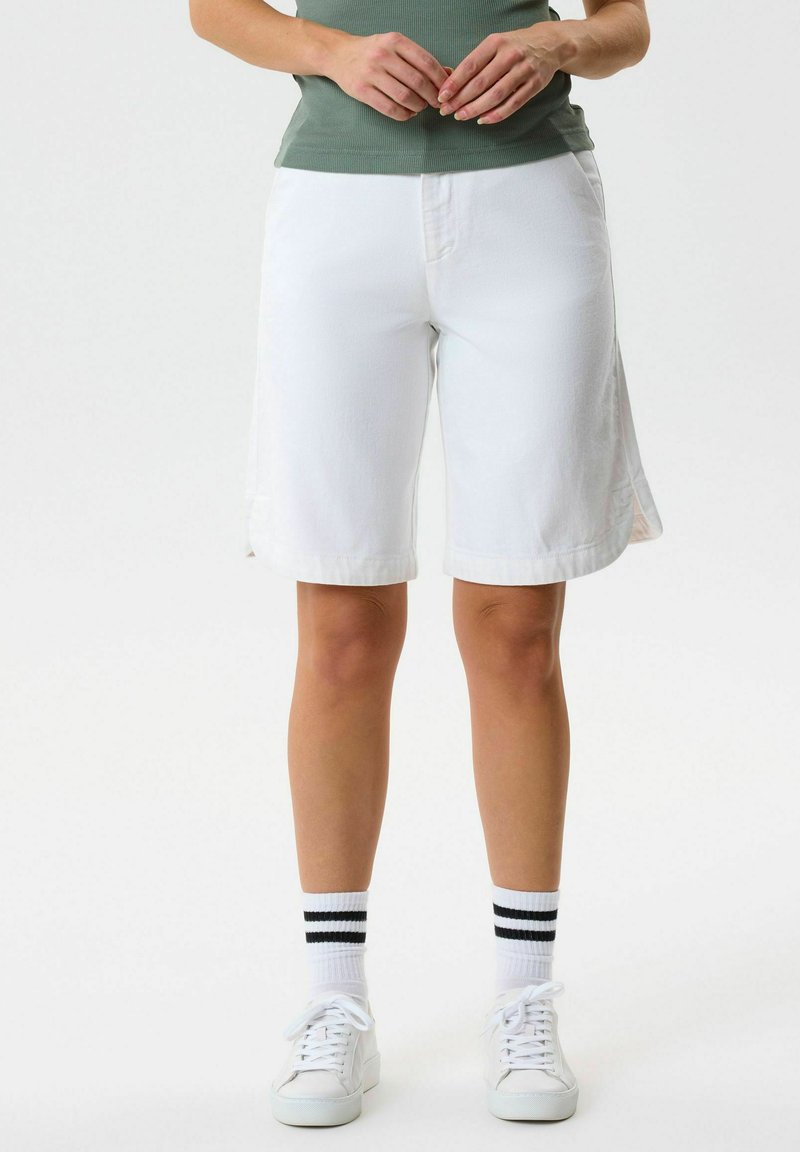 Άτομο που φοράει λευκά σορτς μέχρι το γόνατο, λευκά αθλητικά παπούτσια και λευκές κάλτσες με δύο μαύρες ρίγες, στέκεται μπροστά σε απλό φόντο.