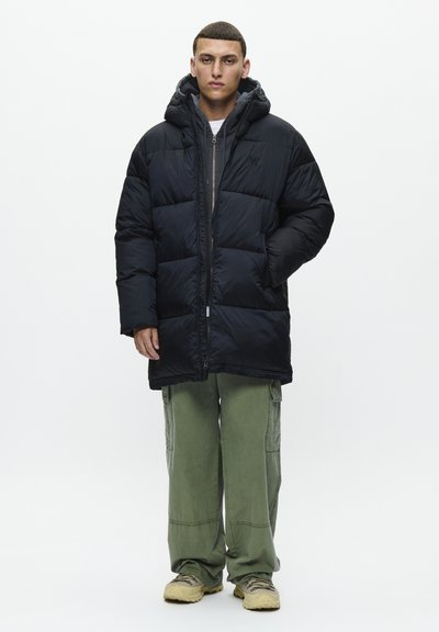 Man wearing black padded coat over a zipped hoodie, green cargo pants, and beige sneakers, standing with hands in pockets on white background.