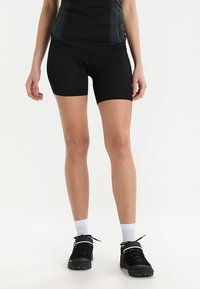 Woman wearing black athletic shorts, black and blue fitted top, white socks, and black sneakers standing against plain white background.