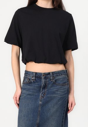 Mujer con una blusa negra de manga corta y corta, y una falda de mezclilla azul de talle alto, mostrada de los hombros a las caderas.