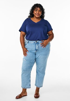 Navy blue v-neck t-shirt, light blue distressed straight-leg jeans, brown flat sandals, medium-length curly hair, neutral backdrop.
