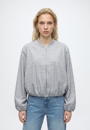 Jonge vrouw met golvend blond haar draagt een grijze geruite jas met rits en een blauwe spijkerbroek, staand tegen een effen witte achtergrond.