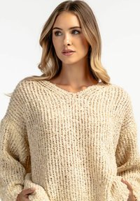 Beżowy sweter wykonany z dzianiny o luźnym kroju, z dekoltem w serek i szerokimi rękawami. Teksturowany materiał ma wzór prążkowany oraz widoczne szwy.