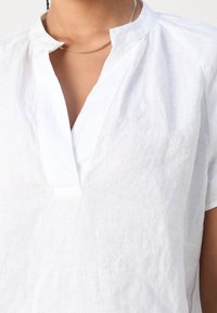 Haut en lin blanc avec un décolleté peu profond, manches courtes et surface texturée, avec des détails froncés près du col.