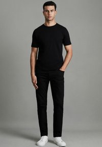 Tricou negru cu guler rotund purtat cu pantaloni negri slim-fit și adidași albi. Modelul stă cu mâinile în buzunare pe un fundal neutru.