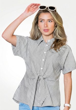 Kobieta o falujących blond włosach nosi szarą i białą koszulę w paski, zapinaną na guziki i związaną w talii, trzymając białe okulary przeciwsłoneczne nad głową.