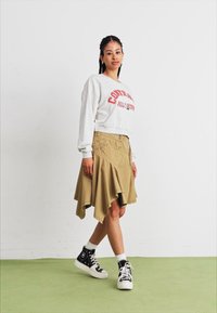 Sudadera gris recortada con texto rojo, falda caqui acampanada con dobladillo asimétrico y zapatillas negras de caña alta con suela blanca. Suelo verde.