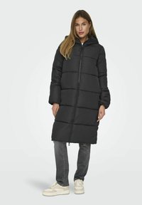 Manteau rembourré noir long avec fermeture éclair à l'avant, capuche et poignets élastiques. Présente un design matelassé avec un ourlet légèrement effilé. Porté par-dessus un jean gris.