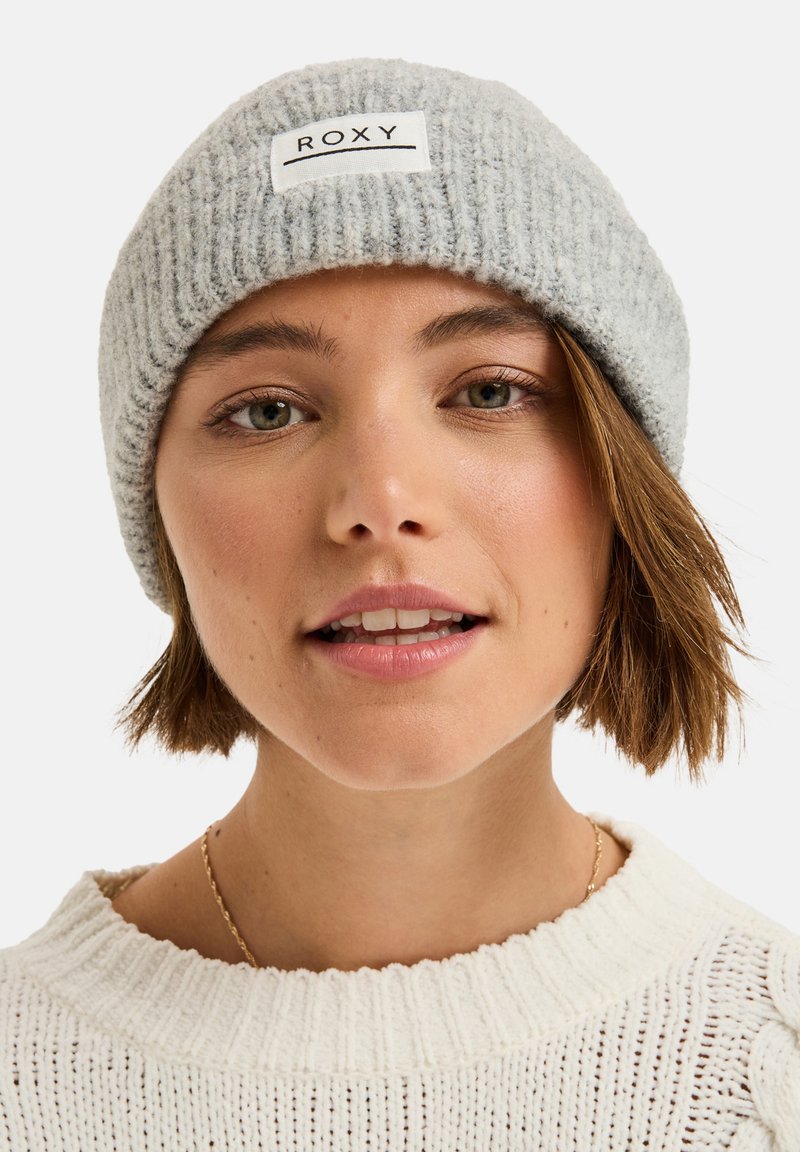 Szara, prążkowana czapka beanie z białą łatą z napisem "ROXY". Miękka faktura, dopasowany krój, noszona pod lekkim kątem na głowie.