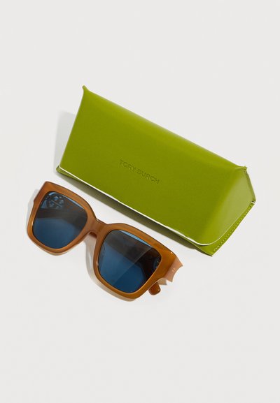 Καφέ τετράγωνα γυαλιά ηλίου με σκούρους φακούς δίπλα σε μια λαχανί σκληρή θήκη Tory Burch πάνω σε λευκή επιφάνεια.