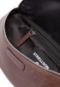 Innenansicht einer braunen Ledertasche mit glatter Textur, Reißverschlusstasche und Stofffutter. Strellson-Label am Rand des Fachs sichtbar.
