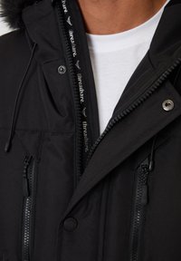 Czarna wodoodporna parka z zapięciem na zamek, dwoma kieszeniami na zamek z przodu oraz kapturem wyłożonym futrem. Posiada oznakowania marki wzdłuż zamka.