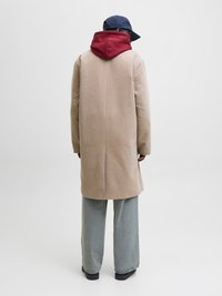 Beige uldfrakke med en bordeaux hætte, sat sammen med lyseblå vide bukser og en marineblå kasket, set fra ryggen mod en neutral baggrund.