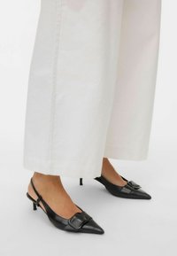 Pies con zapatos de tacón puntiagudo negros tipo slingback con una hebilla grande, combinados con pantalones anchos blancos recortados sobre un fondo liso.