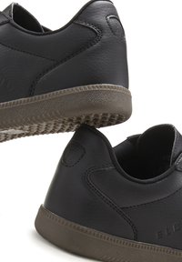 Black leather sneakers feature a low-profile design, textured surface, and contrasting rubber outsole with triangular patterns. Brand logo on the heel.