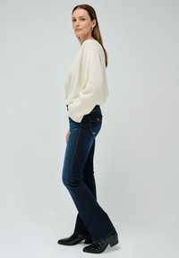 Suéter blanco holgado, jeans de campana azul oscuro y botas de tacón negras. La mujer está de perfil contra un fondo claro.