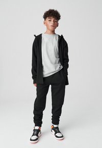 Felpa nera con zip sopra una t-shirt grigio chiaro, abbinata a pantaloni da ginnastica neri e scarpe da ginnastica bianche con dettagli arancioni. Look casual.